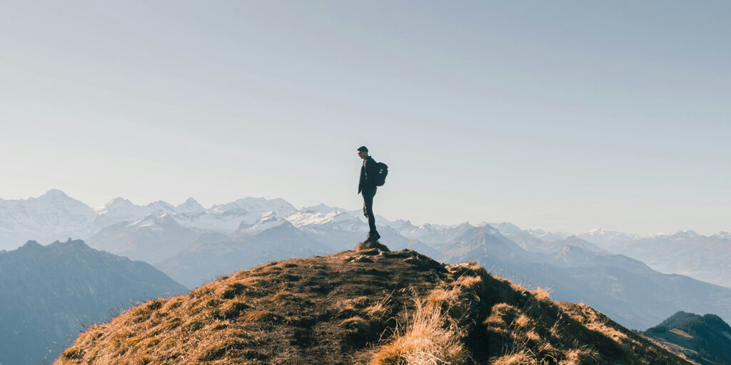 far away view of a person standing on the top of a peak with mountains in the background behind them as they look off to the side, innovative portfolios perspectives