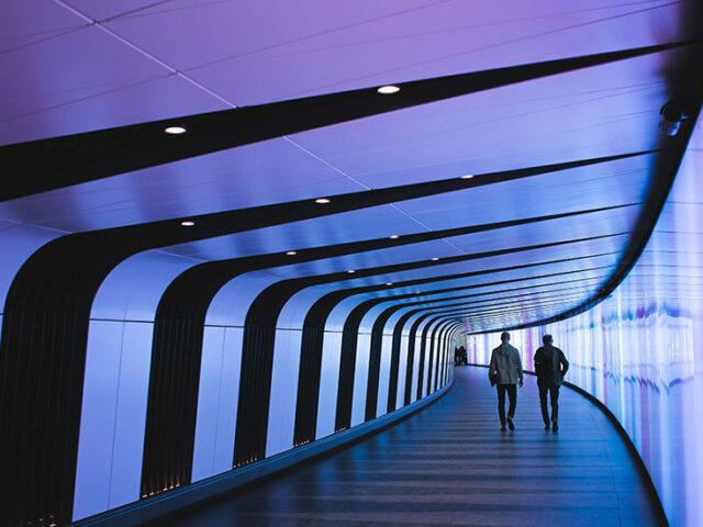 the backs of two people walking down a curved hallway that is illuminated with purple light, innovative portfolios perspectives